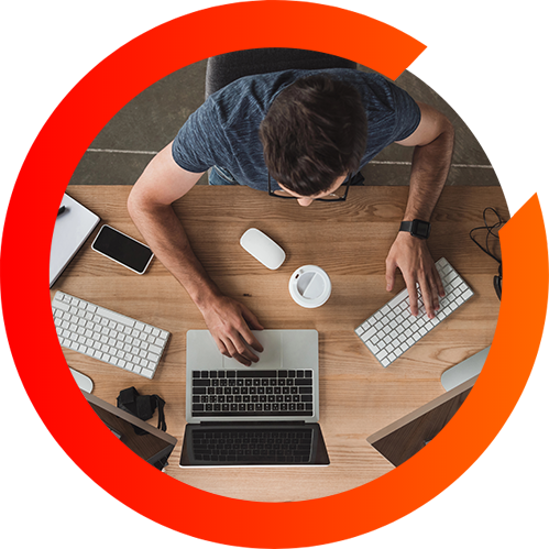 A top down view of a man working hard on a desktop computer and a laptop in a three quarter orange circle.